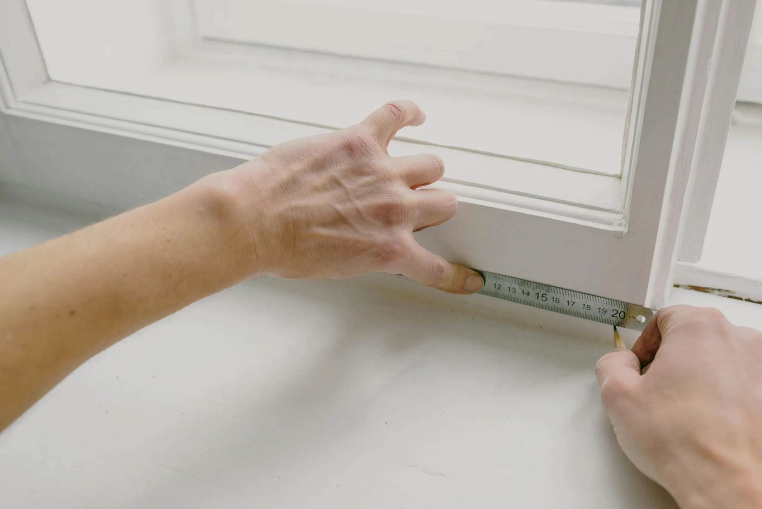 The image shows a close-up view of two hands measuring a white window frame or windowsill using a metal ruler. One hand is pressing the ruler against the edge of the frame while the other hand holds a pencil marking a measurement at around 20 cm. The background and surface are plain and also white. The lighting is natural, coming from the window above, creating soft shadows.