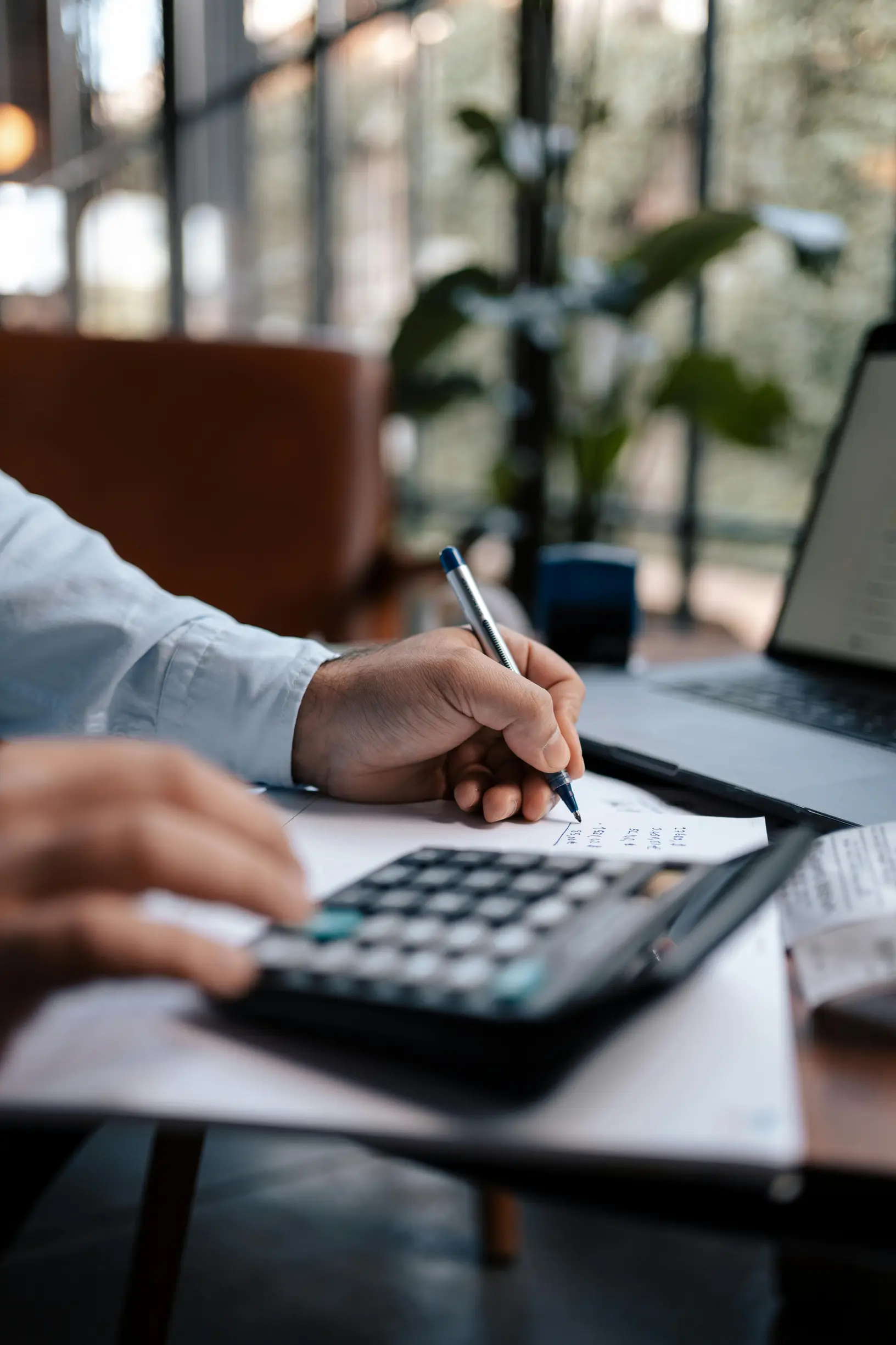 The image shows a close-up view of a person's hands working at a desk. One hand is holding a pen, writing on a piece of paper, while the other is near a calculator. The person is wearing a light blue long-sleeved shirt. There is a laptop with a blank or dimly lit screen in the background, along with a blurred indoor setting featuring a large window and a green plant. The environment suggests an indoor workspace, possibly an office or home office, with natural light coming through the window. The camera angl
