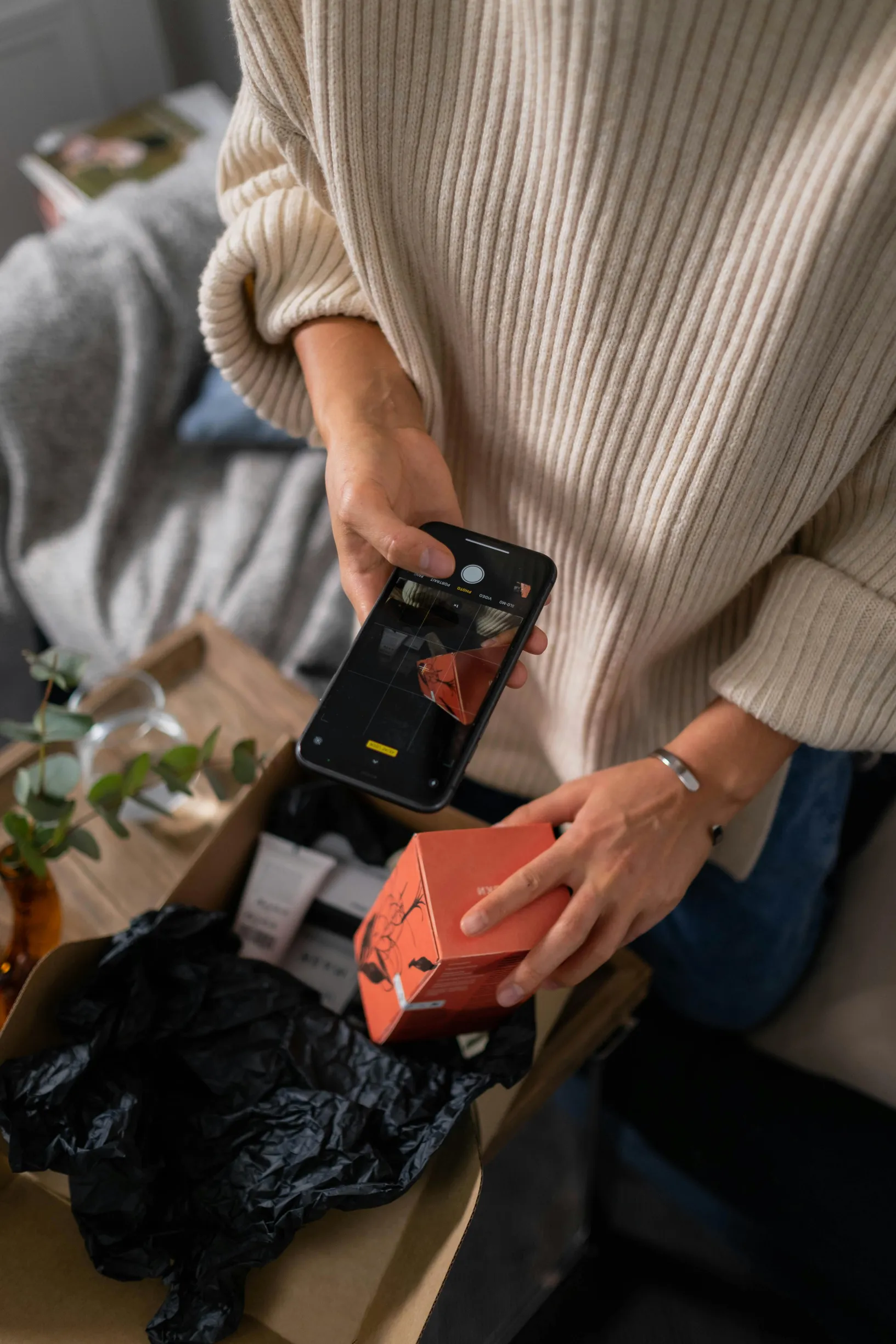 The image shows a person from the torso down, wearing a beige ribbed sweater with sleeves rolled up. They are holding a black smartphone in one hand and an orange rectangular box in the other. The person is seated, and in front of them is an open cardboard box containing a black crumpled fabric. The background shows a soft, gray textile and some leafy green plant elements. The lighting is natural and soft, and the framing is a medium close-up focusing on the hands and objects.