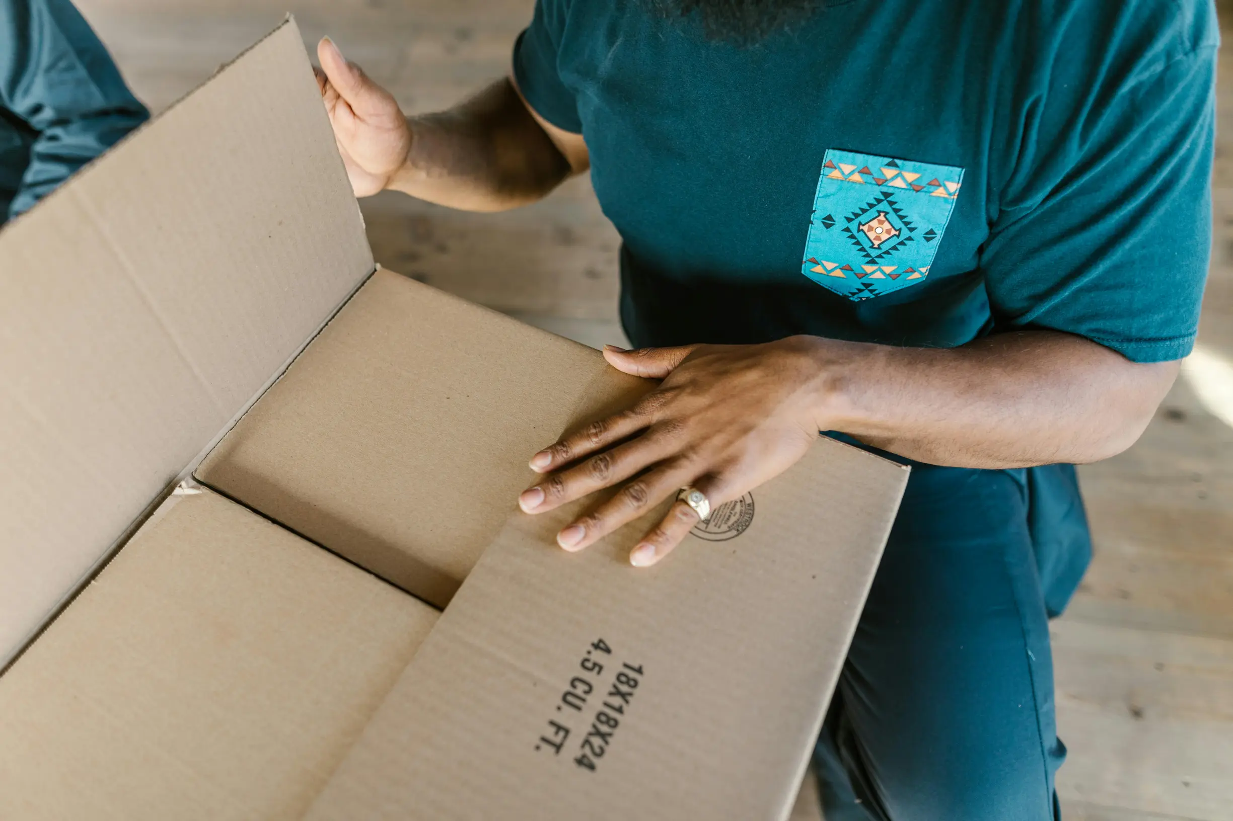 The image shows a person opening a large cardboard box indoors on a wooden floor. The person is visible from the shoulders to the hands, wearing a blue t-shirt with a patterned turquoise pocket and dark pants. The person has a medium to dark skin tone and is wearing a ring on the left hand. The box is partially open and empty, oriented diagonally in the frame.