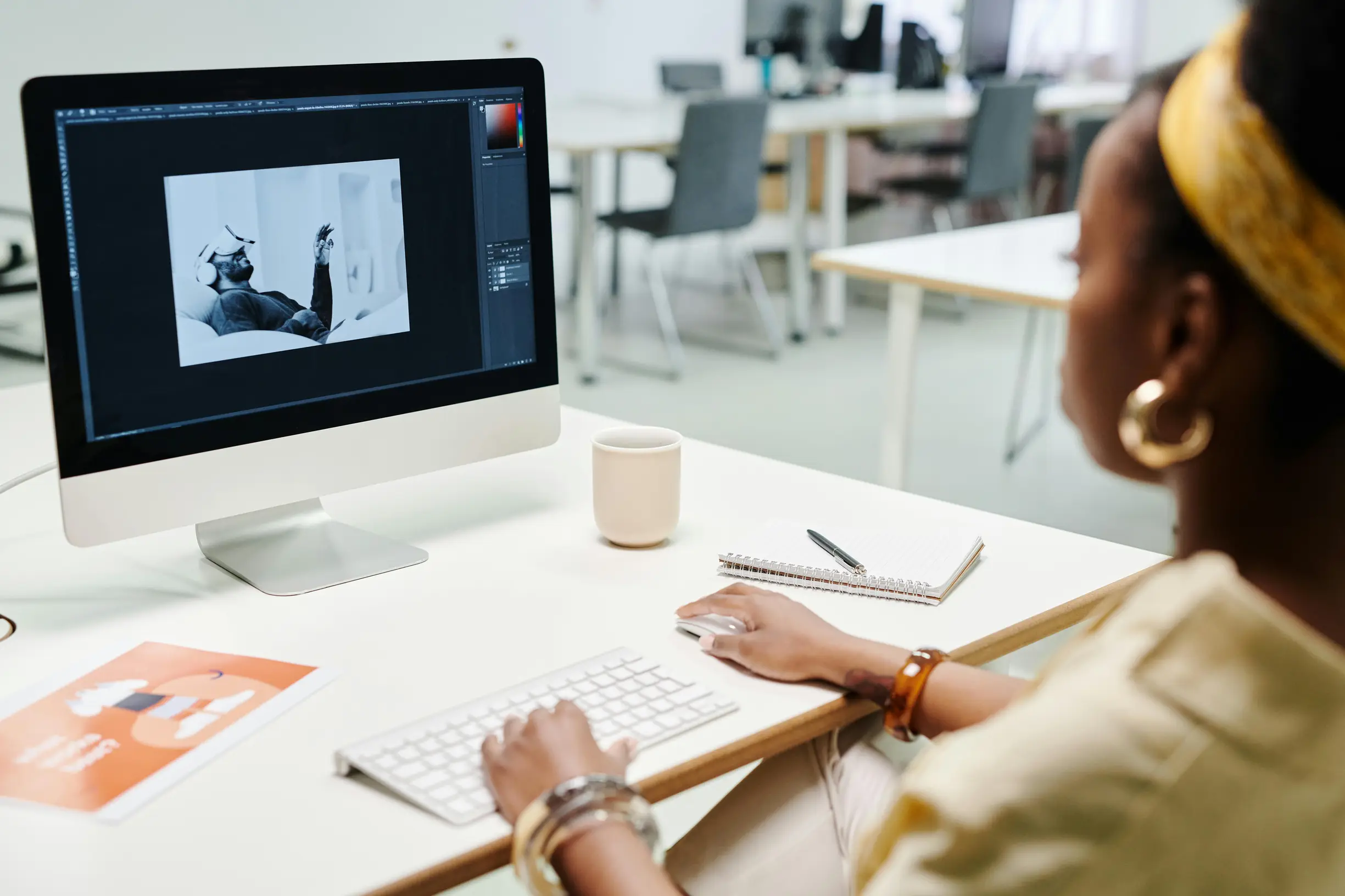 The image shows a person seated at a desk working on a computer. The individual appears to be a woman with dark skin and short hair, wearing a yellow headband, earrings, and bracelets. The computer screen displays a black and white photo editing application with an image of a person performing a handstand. The setting is an office environment with other desks and chairs visible in the background. The desk has a white keyboard, mouse, a closed notebook with a pen on top, a light-colored mug, and a red bookle