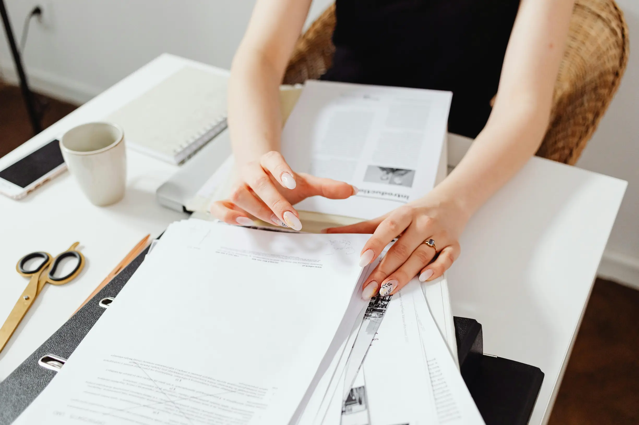 The image shows a person sitting at a white table, organizing or flipping through a stack of documents or papers. Only the person's arms and hands are visible; they have light skin and manicured nails with a light-colored polish. They wear a ring on one finger and a sleeveless black top. On the table are various items including a pair of scissors, a white coffee mug, a smartphone, and a closed notebook. The background is minimalistic with a light-colored wall and a wicker chair.