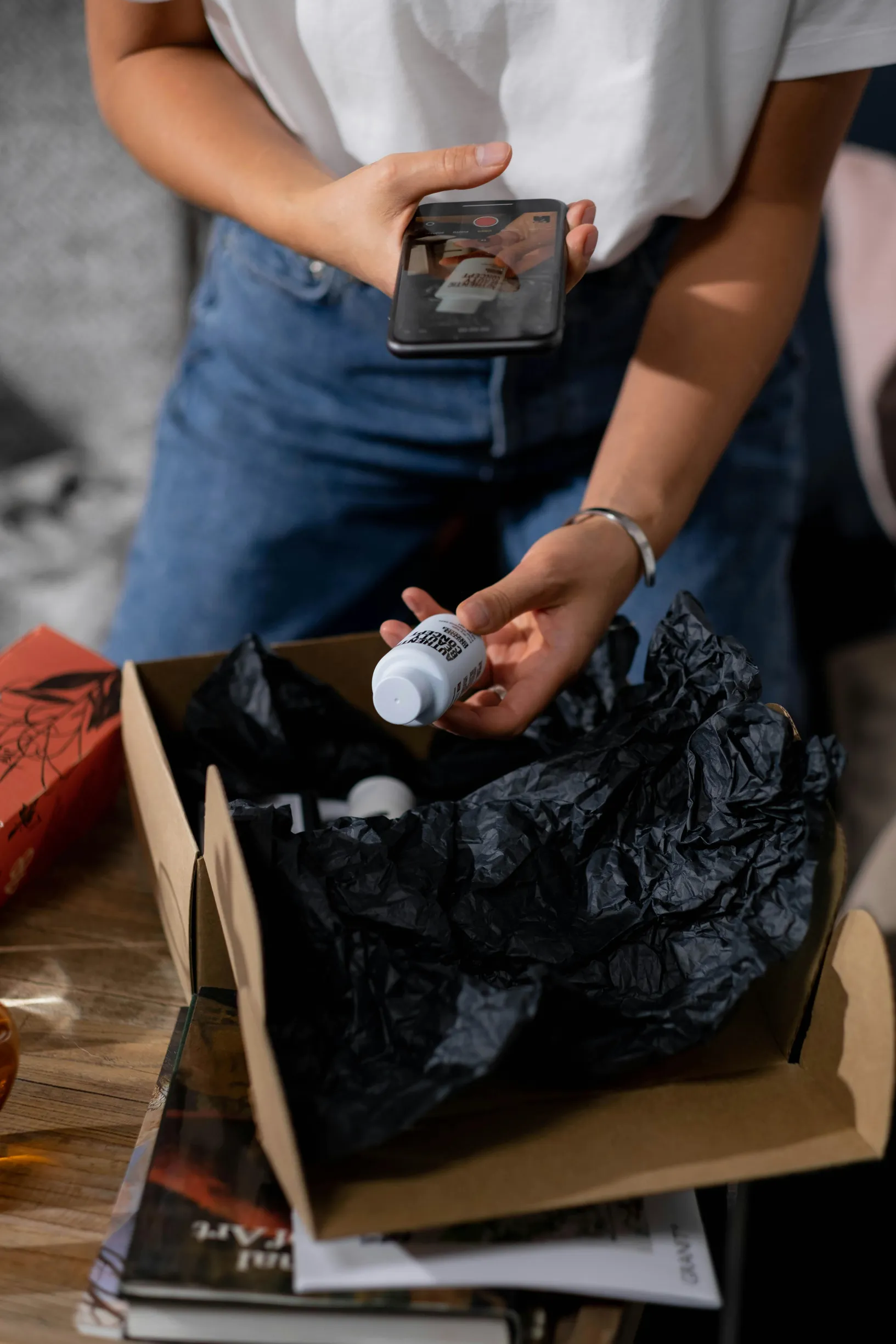 The image shows a person standing beside a table, holding a smartphone in one hand while using the other hand to hold a small white bottle. The person is wearing a white short-sleeve shirt and blue jeans. On the table is an open cardboard box filled with black tissue paper, containing multiple small white bottles. Surrounding the box are some papers or magazines. The scene is lit with soft natural light from the left side and appears to be indoors with a neutral background.
