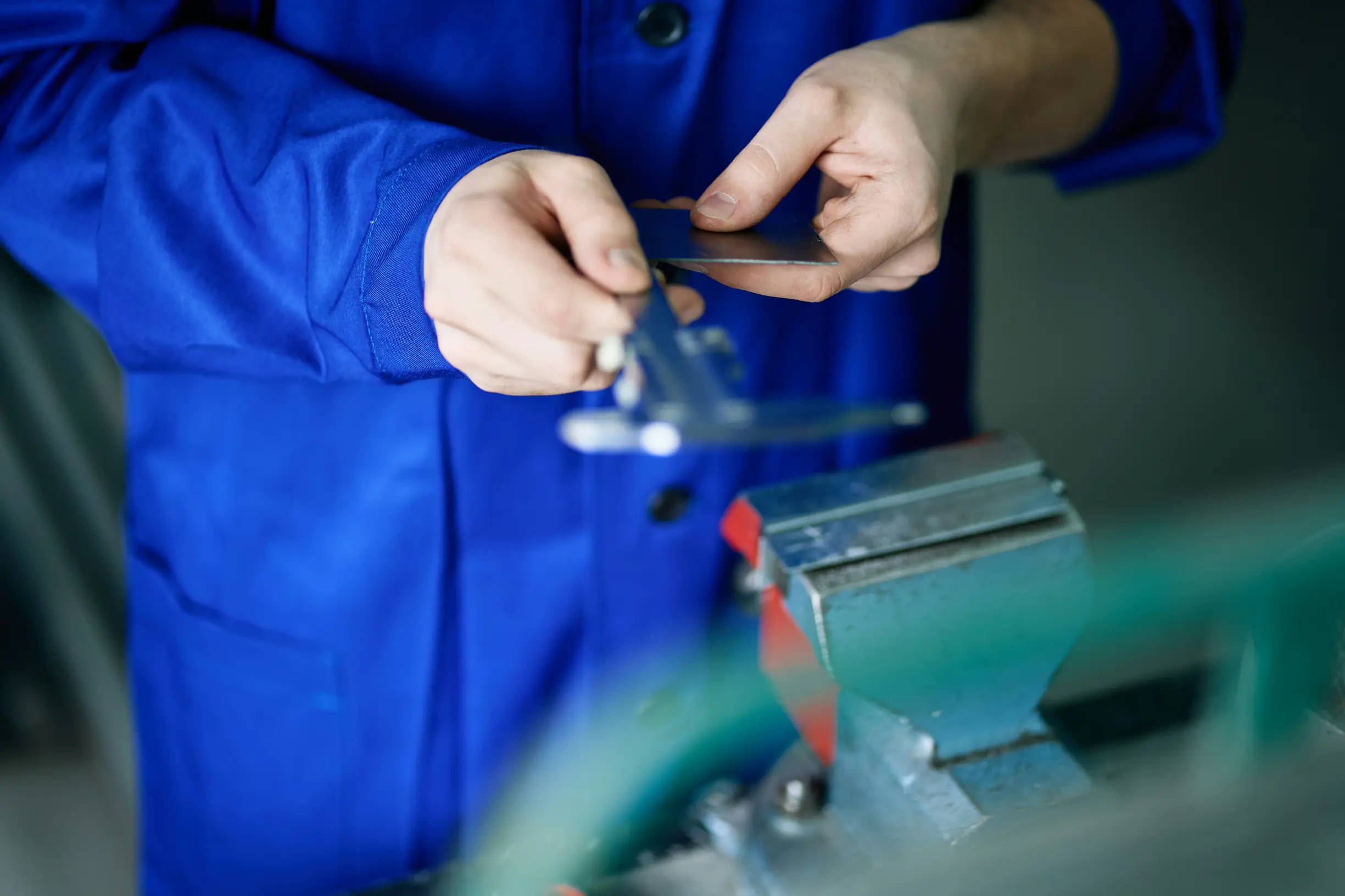 The image shows a person wearing a blue work coat holding a caliper and a small metallic rectangular object. The individual is using the caliper to measure the object's dimensions. A metal vise clamp is visible in the foreground on the right side, out of focus. The background appears to be an industrial or workshop setting with blurred equipment.