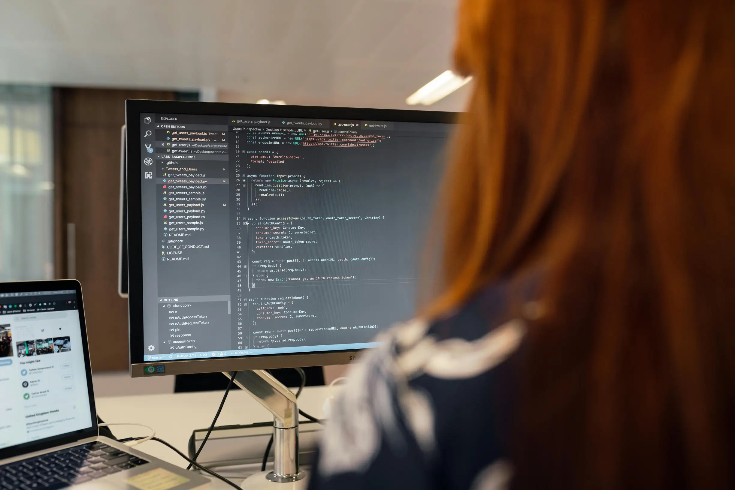 The image shows a person with long reddish hair viewed from behind, sitting at a desk with two screens in front of them. The larger screen displays lines of computer code in a dark-themed text editor, while the smaller laptop screen to the left shows a social media or website interface. The setting appears to be an indoor office environment with blurred background elements. The lighting is natural or evenly distributed. The camera angle is over-the-shoulder, focusing on the computer screens and the back of 