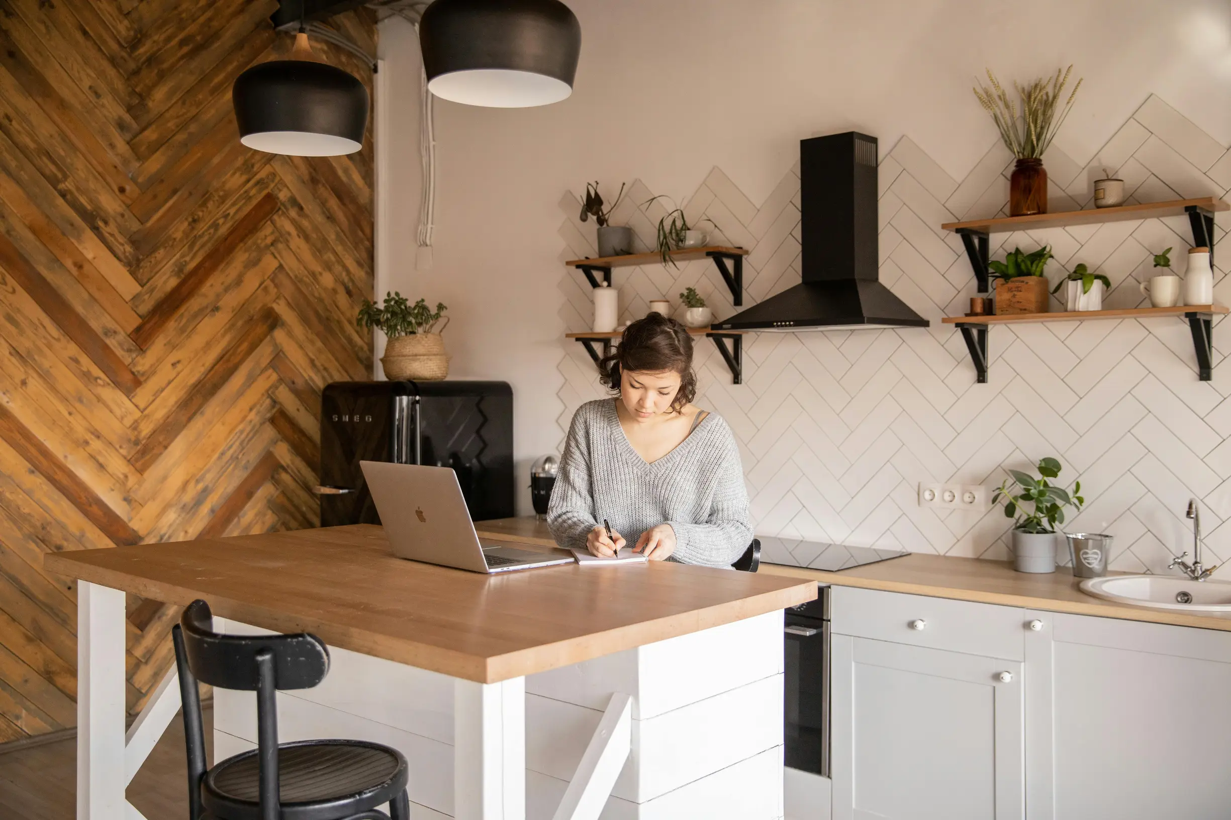 The image shows a young woman sitting at a wooden kitchen island in a modern kitchen. She is writing in a notebook with a pen, with a laptop open in front of her on the island. The kitchen has light-colored cabinetry, a black range hood, white tile backsplash in a diamond pattern, and wooden shelves with small plants and decorative items. The wood-paneled wall on the left has a herringbone pattern. Pendant lights hang from the ceiling above the island. The overall lighting is natural and warm, and the camer