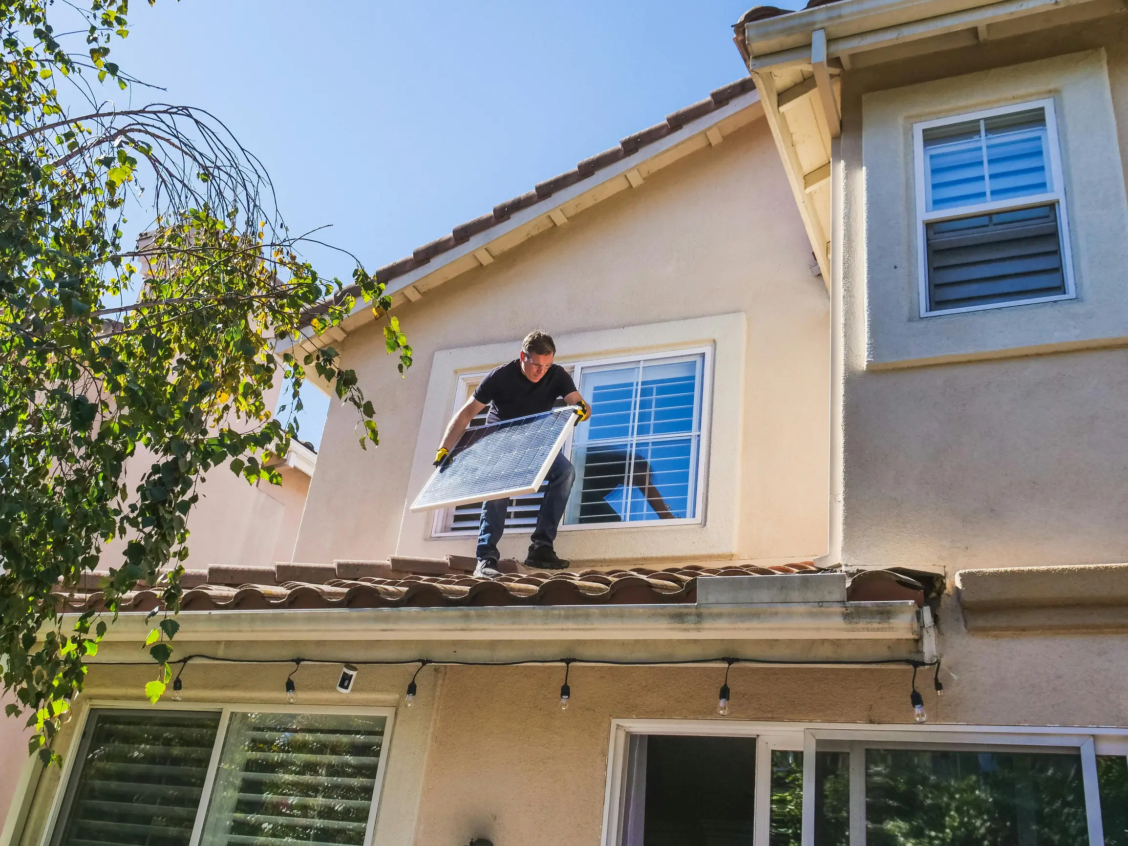 The image shows one person standing on the roof of a two-story residential house. The person appears to be holding a rectangular object, likely a solar panel, while positioned near a window on the upper floor. The house has a beige exterior with white trim and a tile roof. There is a tree with green leaves visible on the left side of the image. The sky is clear and blue, suggesting sunny weather. The camera angle is at eye level from a moderate distance, capturing both the upper portion of the house and par