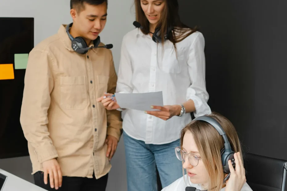 The image shows three people in a modern office setting. Two individuals are standing by a wall with contrasting black and white colors; one is holding a document that both are looking at. Another person is sitting at a desk with a laptop, wearing a headset, and appears to be working or participating in a call. The seated individual also has dyed blonde hair and glasses. The workspace includes a desk with a laptop, disposable coffee cup, papers, and sticky notes on the wall.