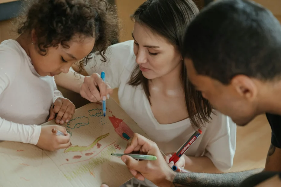 The image shows three people sitting closely together, engaging in a drawing activity on a piece of brown paper. A young girl with curly hair, wearing a white long-sleeve shirt, is on the left. Next to her is a young woman with straight brown hair, also wearing a white top, and on the right is a man with short hair, wearing a black shirt. They are all holding colored markers and focused on coloring and drawing various shapes and lines on the paper.
