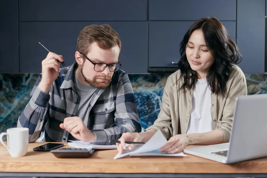 The image shows two adults seated at a wooden table indoors, possibly in a kitchen or modern office space. The man on the left, wearing glasses and a checkered shirt over a gray t-shirt, is holding a pen and focused on a notebook. The woman on the right, with long dark hair and wearing a beige cardigan over a white shirt, is looking down at documents she holds, adjacent to a silver laptop. The background features dark cabinetry and a blue marble backsplash. The lighting is soft and even, suggesting natural