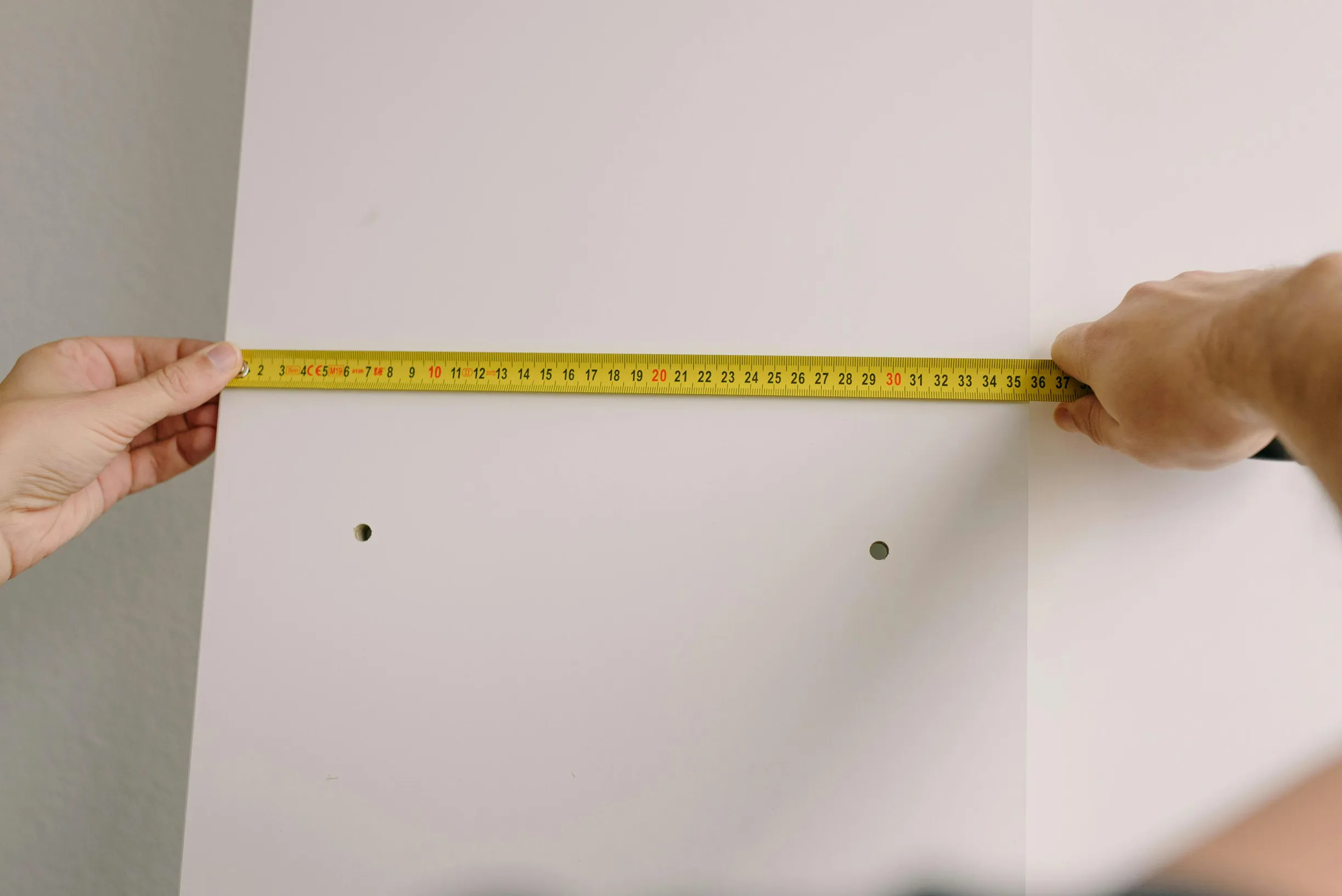 The image shows two hands holding a yellow measuring tape stretched horizontally across a white wall. The measuring tape displays numbers in centimeters, and it extends from left to right, indicating a length of about 30 cm. The wall has two visible holes evenly spaced between the hands, suggesting previous mounting points or drill holes.