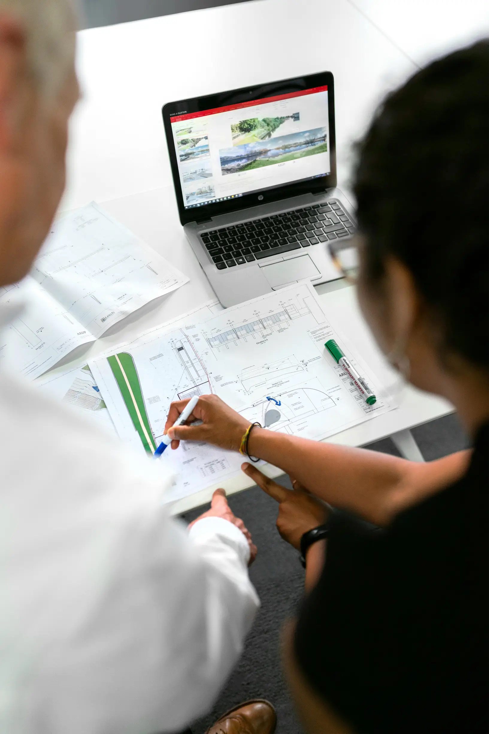 The image shows two people sitting at a desk viewed from behind and slightly above. One person has gray hair and is wearing a white shirt, while the other has dark hair tied back and wears a black top. They are looking at an open laptop displaying a webpage with images and text. In front of them on the desk are architectural or engineering documents, a pen, and a green highlighter, which the darker-haired person is holding. The setting appears to be an indoor office environment with a white desk surface and
