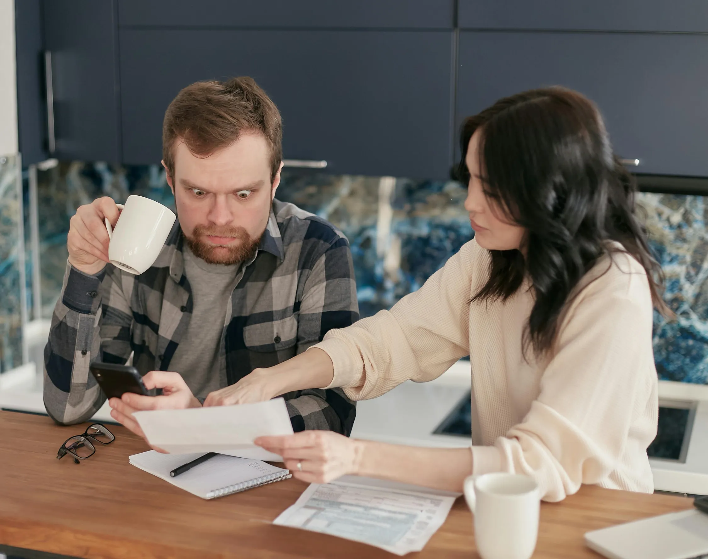 Two adults sit at a kitchen counter reviewing documents. The man on the left wears a grey and black plaid shirt over a t-shirt, holds a mug in his right hand, and looks at a phone with a concerned expression. The woman on the right, with long dark hair and a cream-colored sweater, points and gestures toward the documents spread on the counter. The kitchen backdrop includes dark cabinets and a blue and grey marble backsplash. There are two white mugs, paperwork, a pen, and a notebook on the counter.