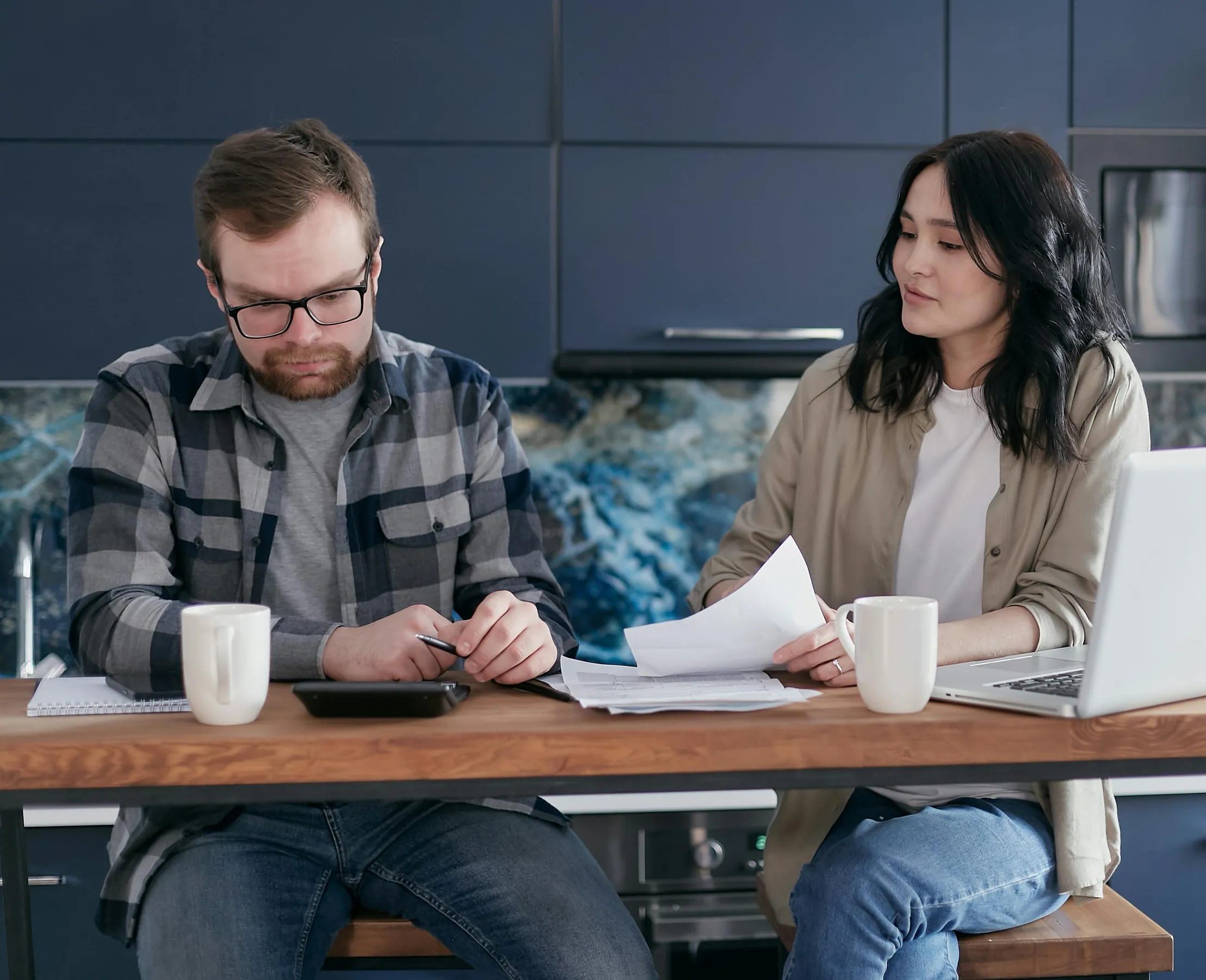 Two people are seated at a wooden table in a modern kitchen setting. The person on the left is a man with light skin and short brown hair, wearing glasses and a blue and gray checkered shirt over a gray t-shirt, looking down and holding a pen near a calculator. To the right, a woman with light skin and long black hair, wearing a beige jacket over a white shirt, holds papers and looks toward the man. On the table are two white mugs, papers, and a silver laptop.
