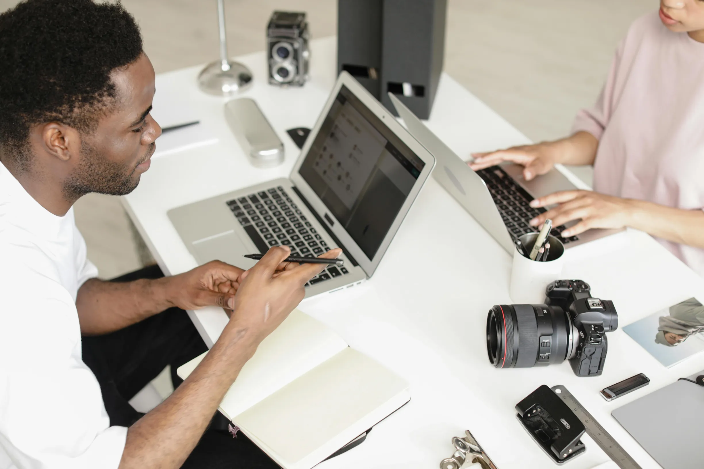 Two people sit at a white table working on laptops. The person on the left is a dark-skinned man wearing a white shirt, holding a pen and facing his open laptop. The person on the right is partially visible, wearing a light pink top and using a laptop as well. On the table are various electronic devices including a DSLR camera with lens attached, a vintage camera, a flash unit, a memory card, and other photography accessories. The environment appears indoors with soft, natural lighting captured from an eye-