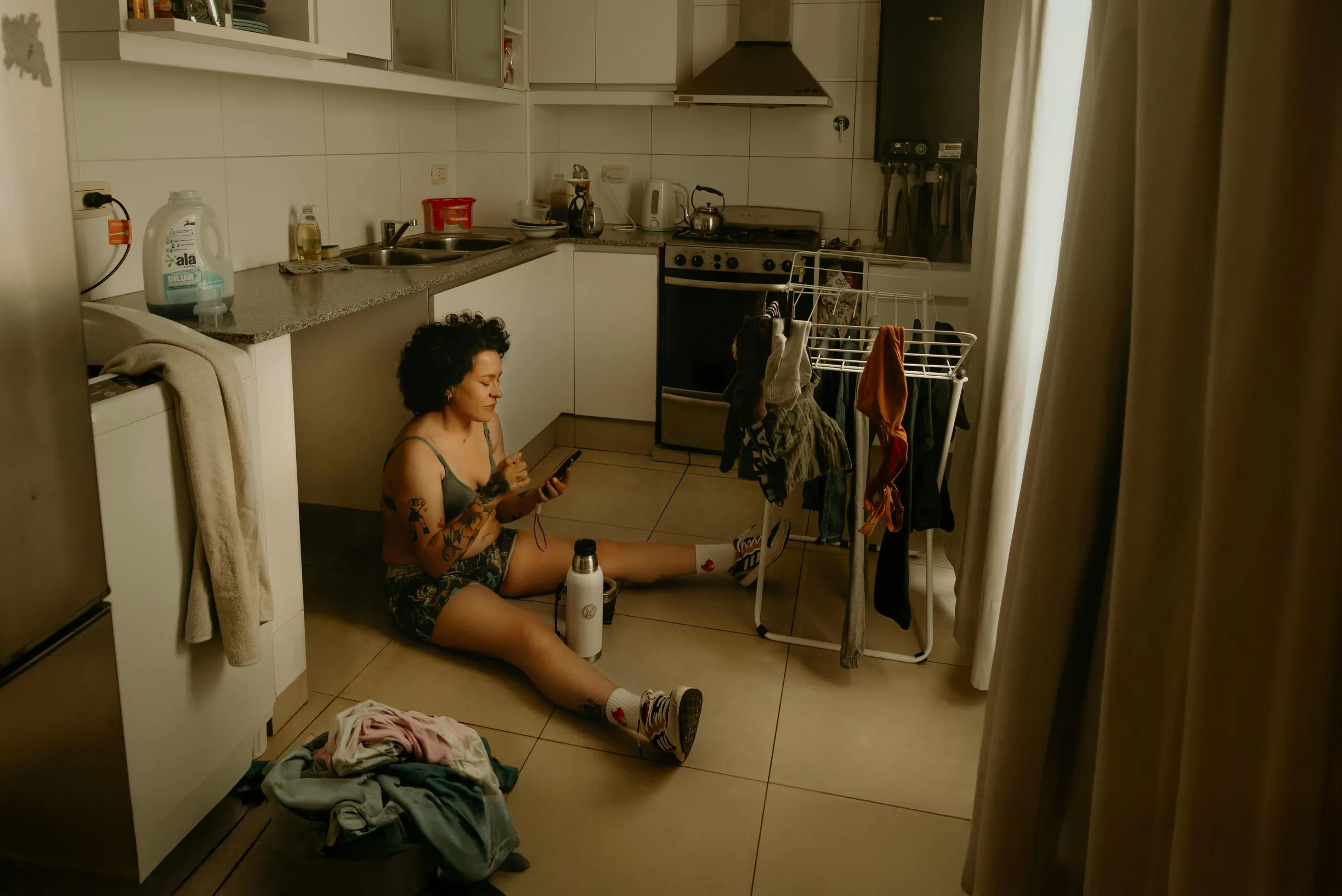 Young woman sitting on kitchen floor using smartphone while doing laundry. Cozy, relaxed indoor setting.