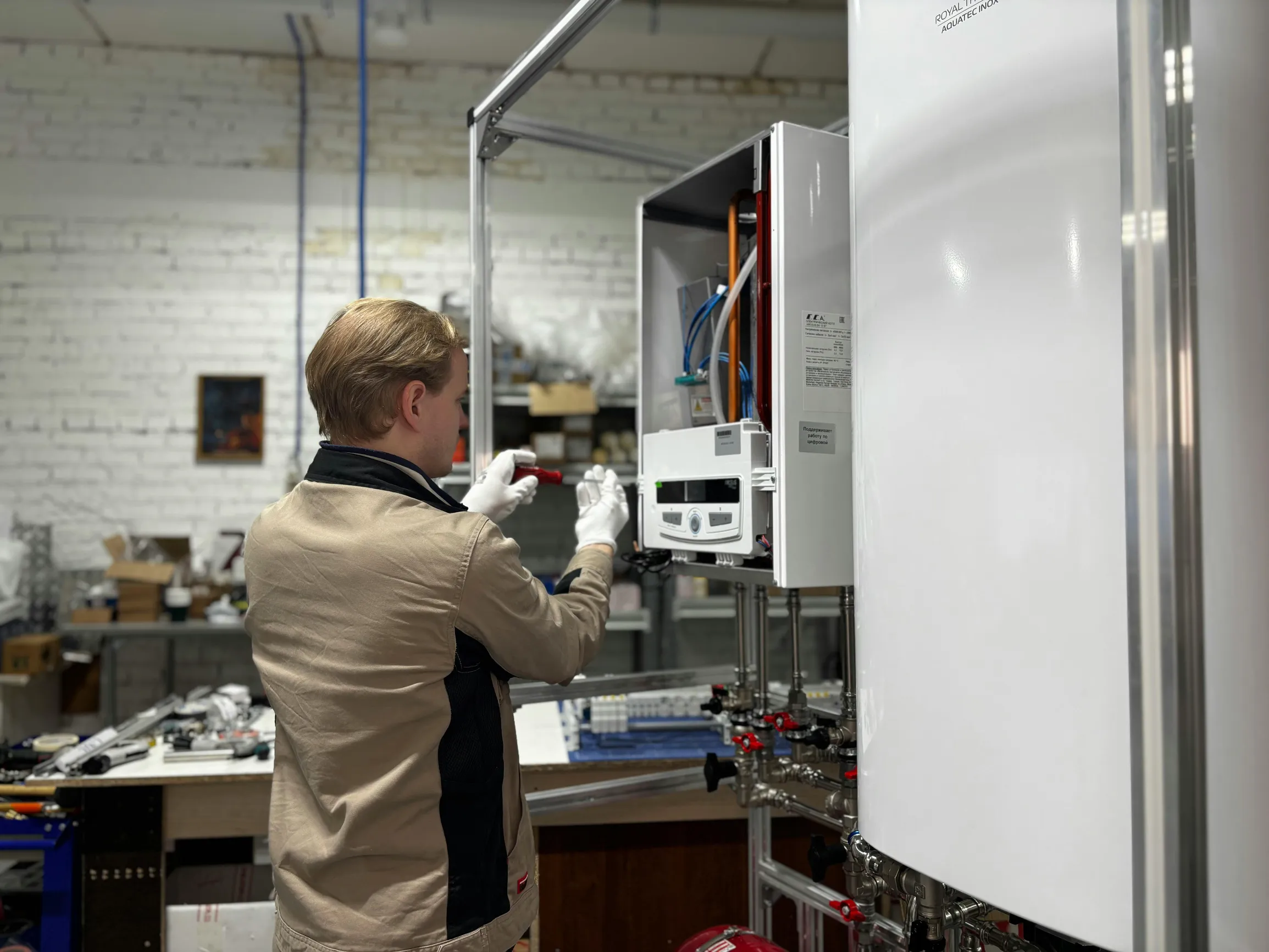 A person is working indoors in a workshop or industrial setting. The individual is standing next to an open white boiler or heating unit, inspecting or repairing its internal components. The person is wearing a beige work jacket and white gloves. The background shows a cluttered workspace with shelves, boxes, and tools, and a white brick wall. The scene is lit with artificial overhead lighting and viewed from an eye-level angle with a medium depth of field.