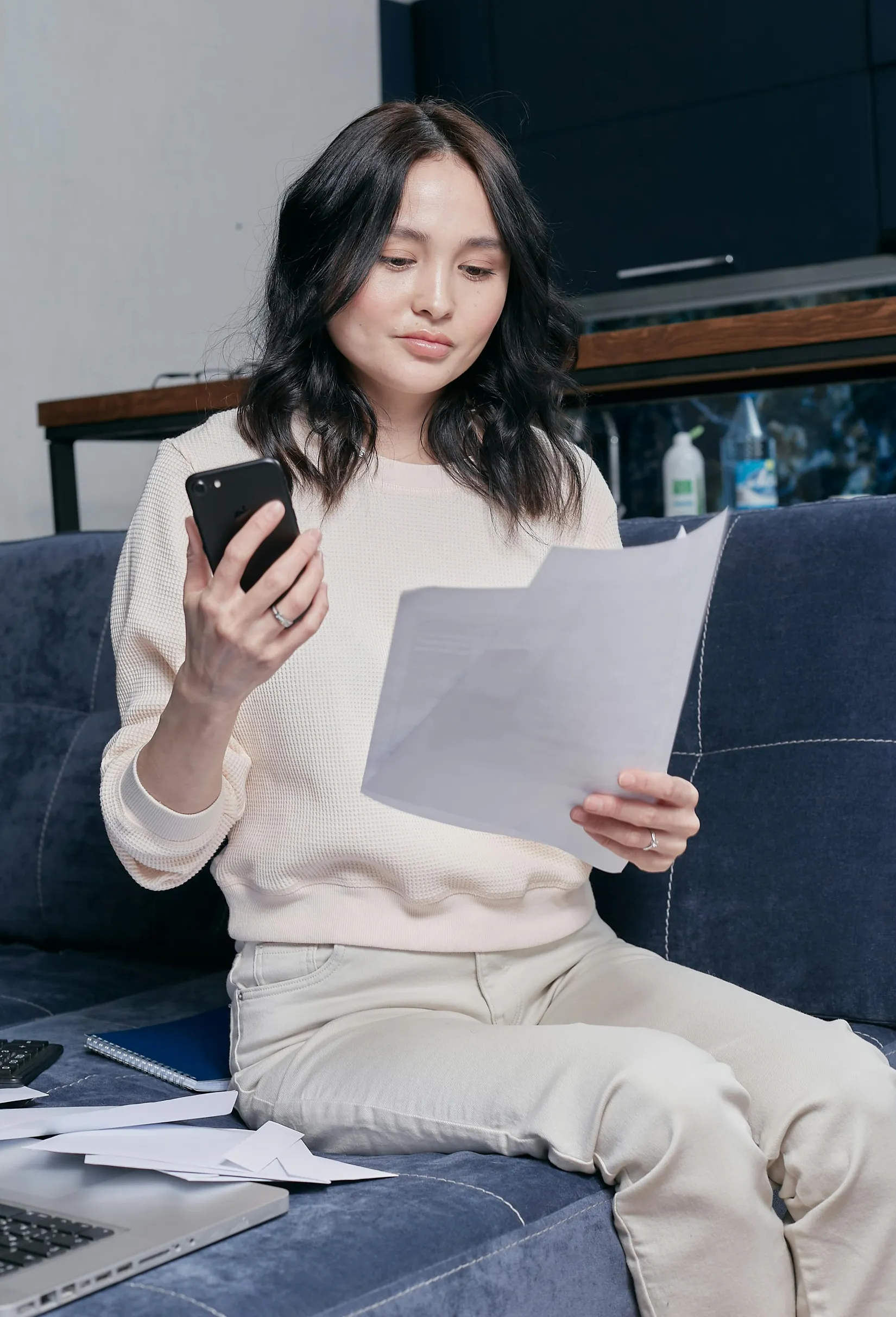 A young woman with shoulder-length dark hair sits on a blue couch. She is wearing a light-colored sweatshirt and cream-colored pants. The woman holds a smartphone in one hand and a piece of paper in the other, looking intently at the paper. Additional papers are scattered on the couch beside her. Behind her, there is a kitchen counter with bottles and a dark-colored cabinet. The lighting is soft and even, suggesting indoor natural or diffused light. The shot is a medium framing at eye-level angle.