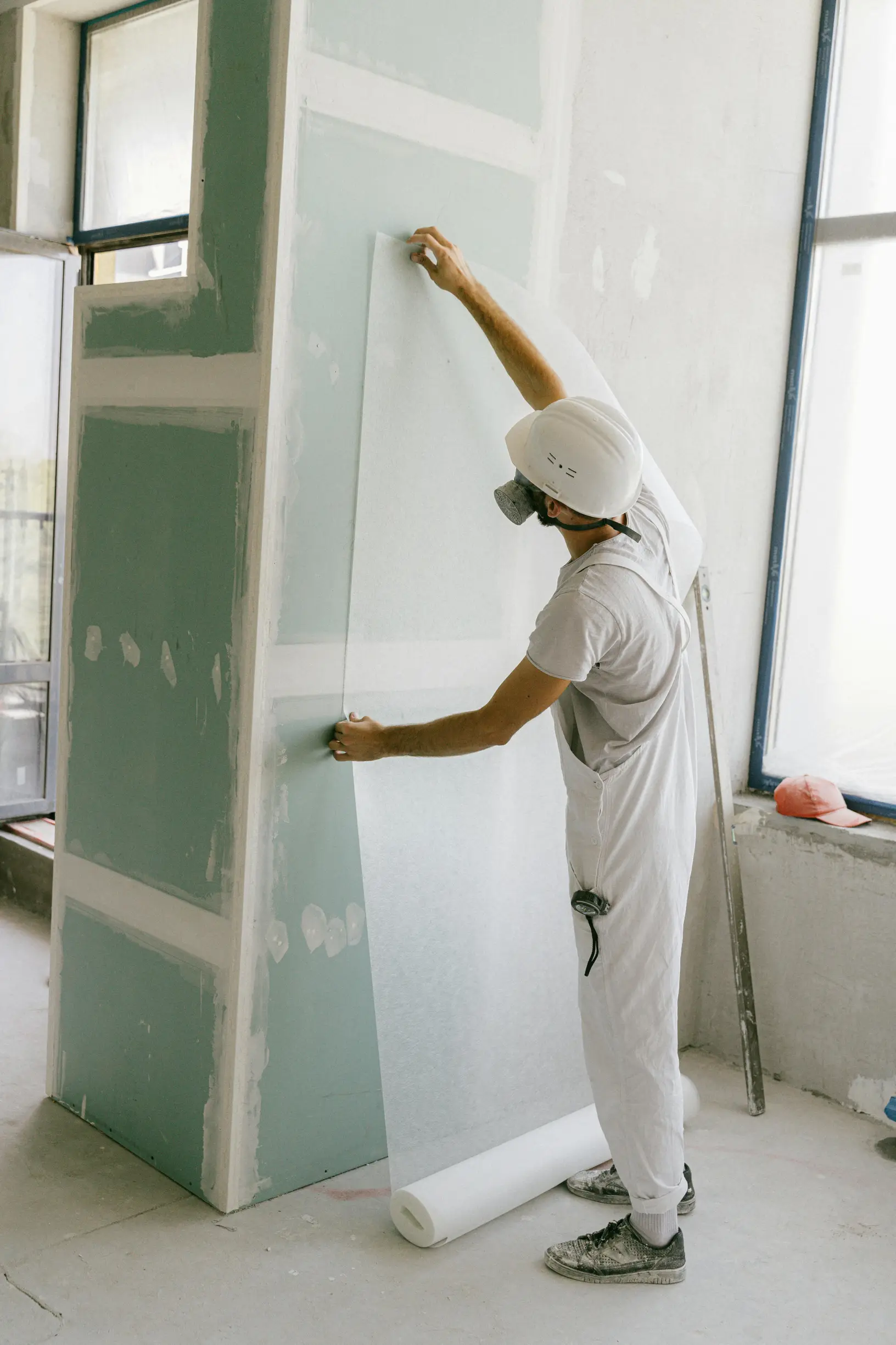 An individual is working indoors on installing or finishing drywall. The person is wearing a white shirt, white overalls, a white protective helmet, and a dust mask. They are standing next to a large, vertical drywall panel, smoothing or applying material to it using tools. The setting appears to be a construction or renovation site with bare floors and large windows allowing natural light into the space. The camera angle is eye-level, capturing the subject in a medium shot from the side, showing the upper