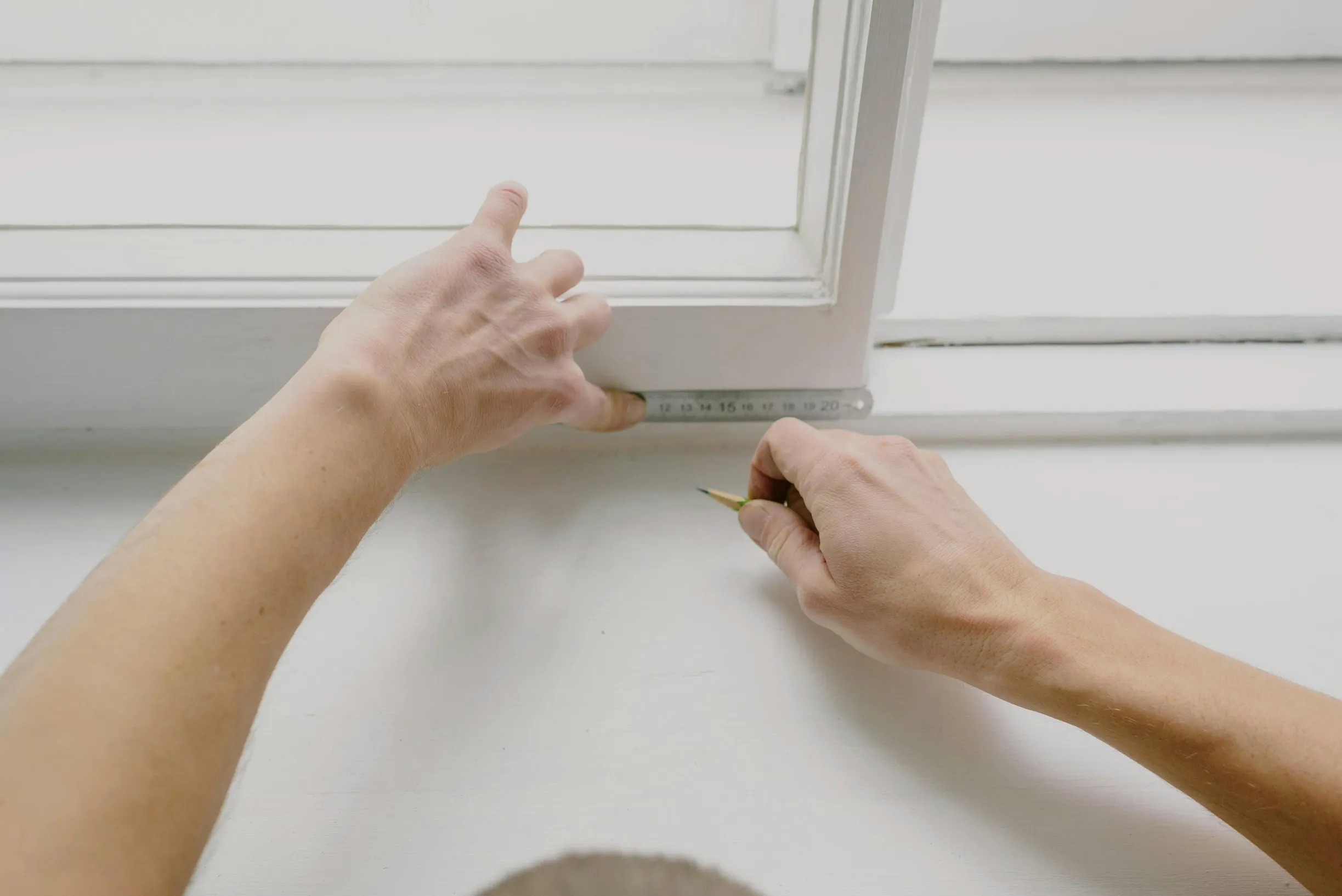 The image depicts two adult hands measuring the width of a white window frame using a metal ruler marked in centimeters. One hand holds the ruler in place at about 20 cm, while the other hand holds a pencil to mark the measurement. The setting appears to be indoors, with the measuring action taking place against a white windowsill under a large white-framed window. The lighting is natural and soft, coming from outside through the window, illuminating the scene evenly. The camera angle is close-up and slight