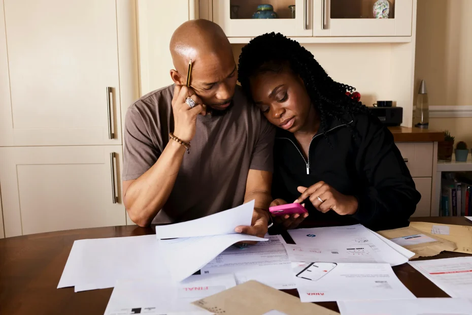 The image shows a man and a woman sitting closely together at a wooden table covered with papers and envelopes. The man is bald, wearing a brownish short-sleeve shirt and holding a pencil to his temple while looking at some documents. The woman has braided hair, is dressed in a black top, and is focused on a pink smartphone she holds, pointing at its screen. Behind them is a cream-colored kitchen setting with cabinets, a countertop, and some household items. The lighting is warm and even, suggesting natural