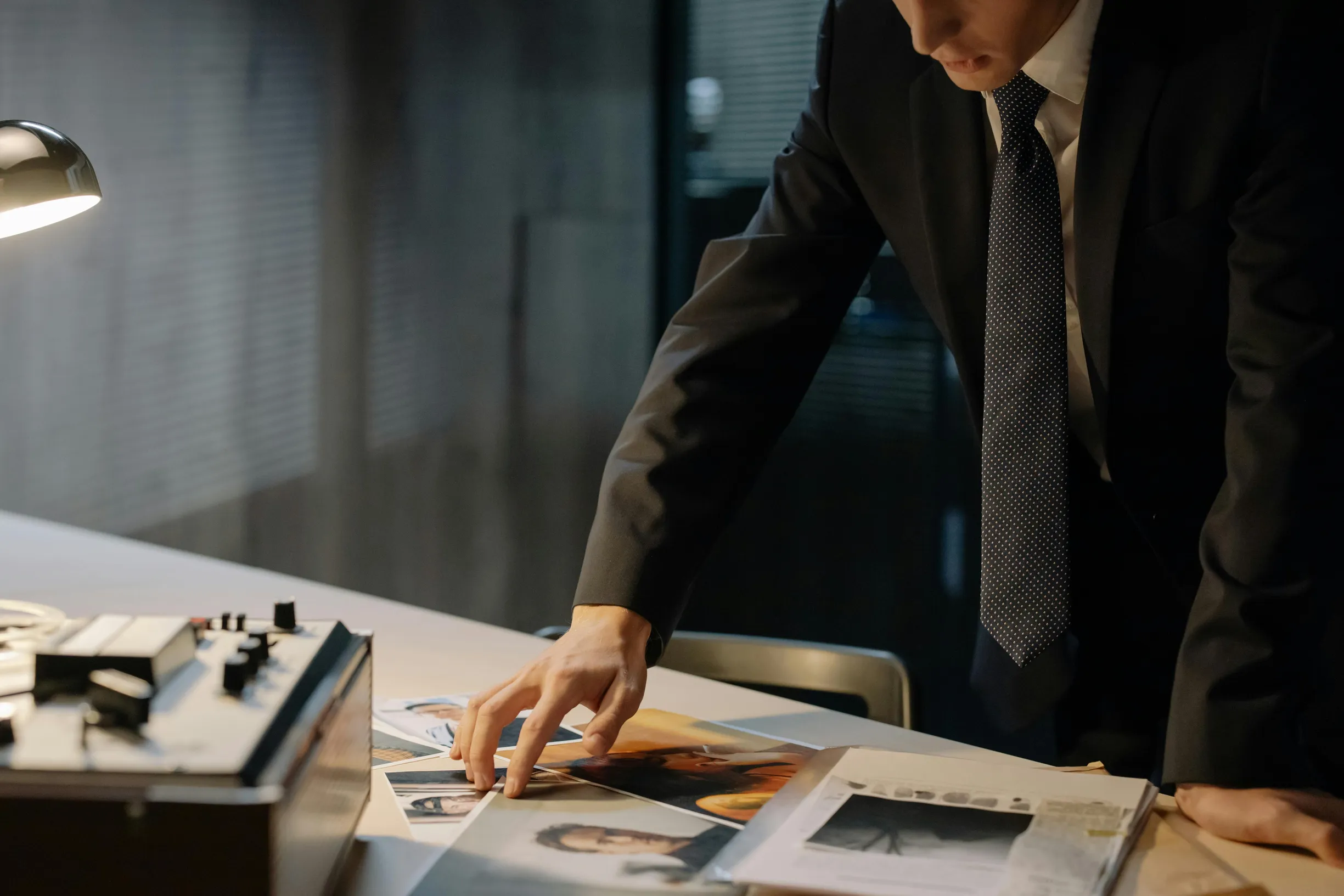 The image shows a man in a dark suit with a white shirt and patterned tie standing and leaning over a desk. He is sorting through printed photographs spread on a white desk surface. There is a lamp on the left side of the desk illuminating the area, and a tape recorder or similar device is partially visible on the desk. The background is dim, suggesting an indoor office or workroom environment with subdued lighting. The camera angle is eye-level, focusing on the upper body of the man and the desk, with a cr