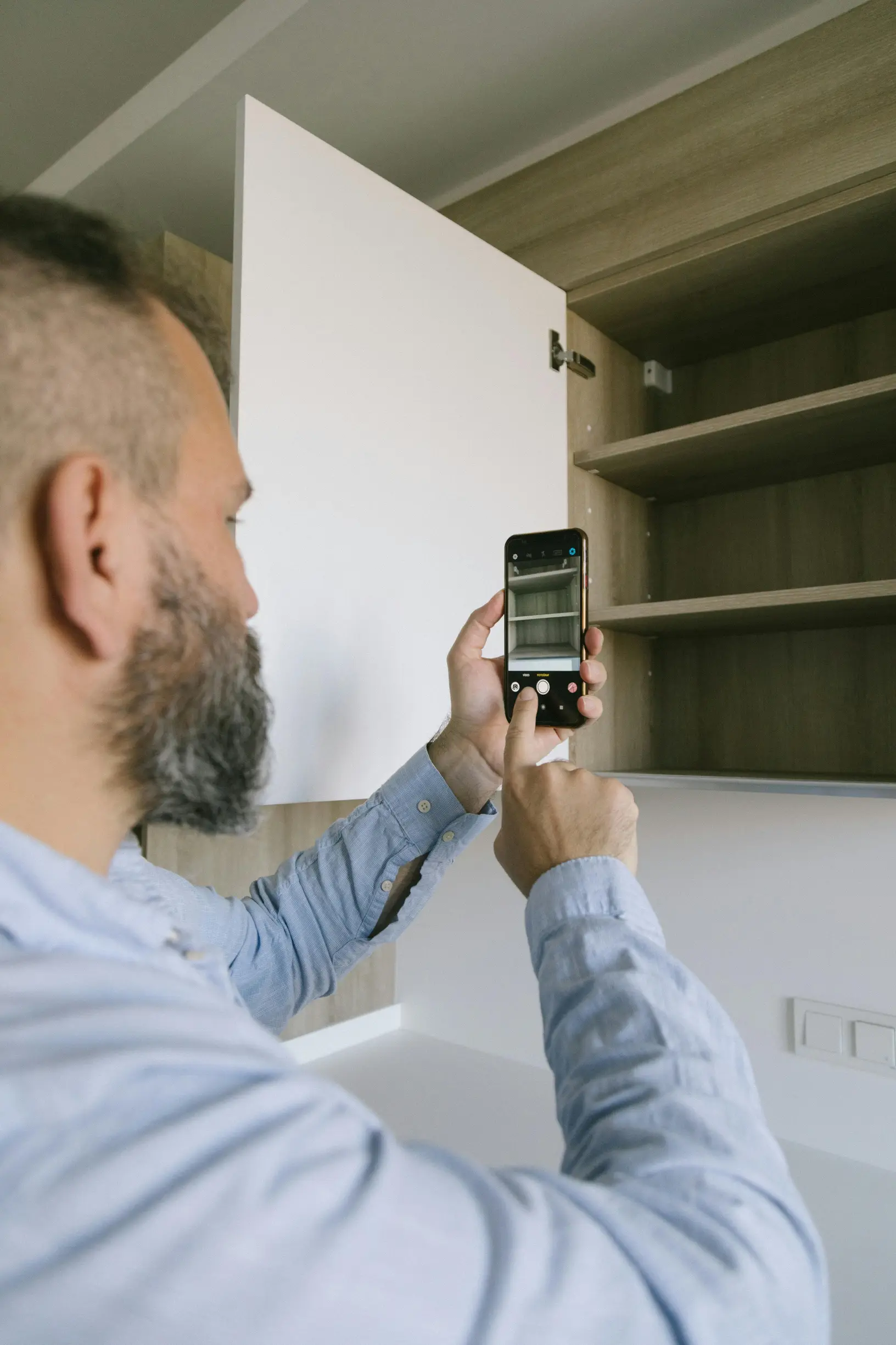 The image shows a man with a beard and short hair viewed from behind and the side, facing an open cabinet in a domestic setting. He is holding a smartphone in one hand and using the other hand to interact with the screen, appearing to take a photo or video inside the cabinet. The cabinet has several empty shelves and a white door, and is mounted on a light-colored wall. The man wears a light blue, long-sleeved shirt. The overall scene is well-lit with diffuse natural or artificial light.