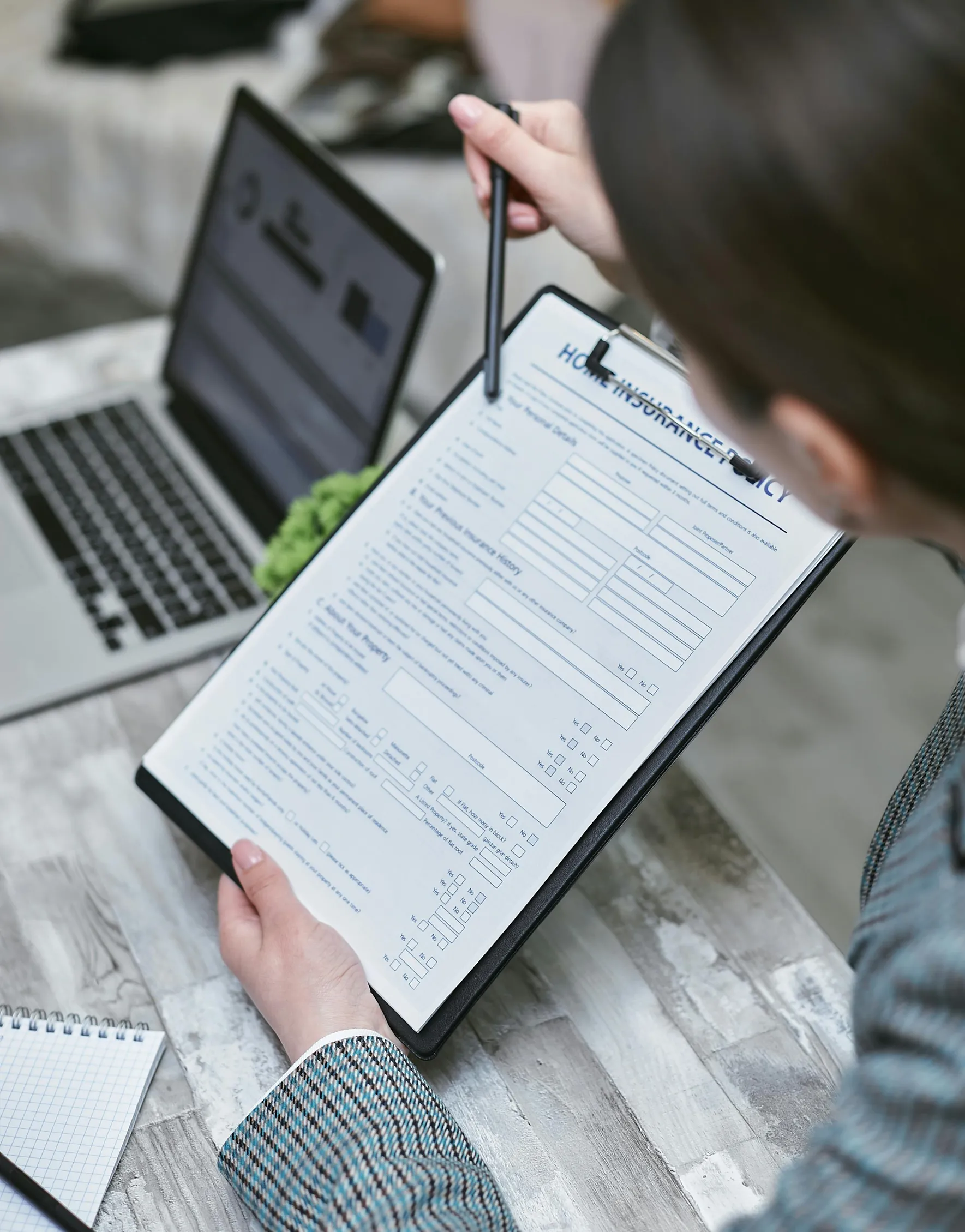 The image shows a person holding a clipboard with a document attached, seemingly reading or writing on it using a pen or pencil. A laptop with an open screen is positioned on a table in front of the person. The table surface appears to be wooden or laminate with a grayish tone. The person's clothing includes a striped shirt with visible cuffs. The image is taken from behind and above the person's shoulder, focusing on the clipboard and the laptop.