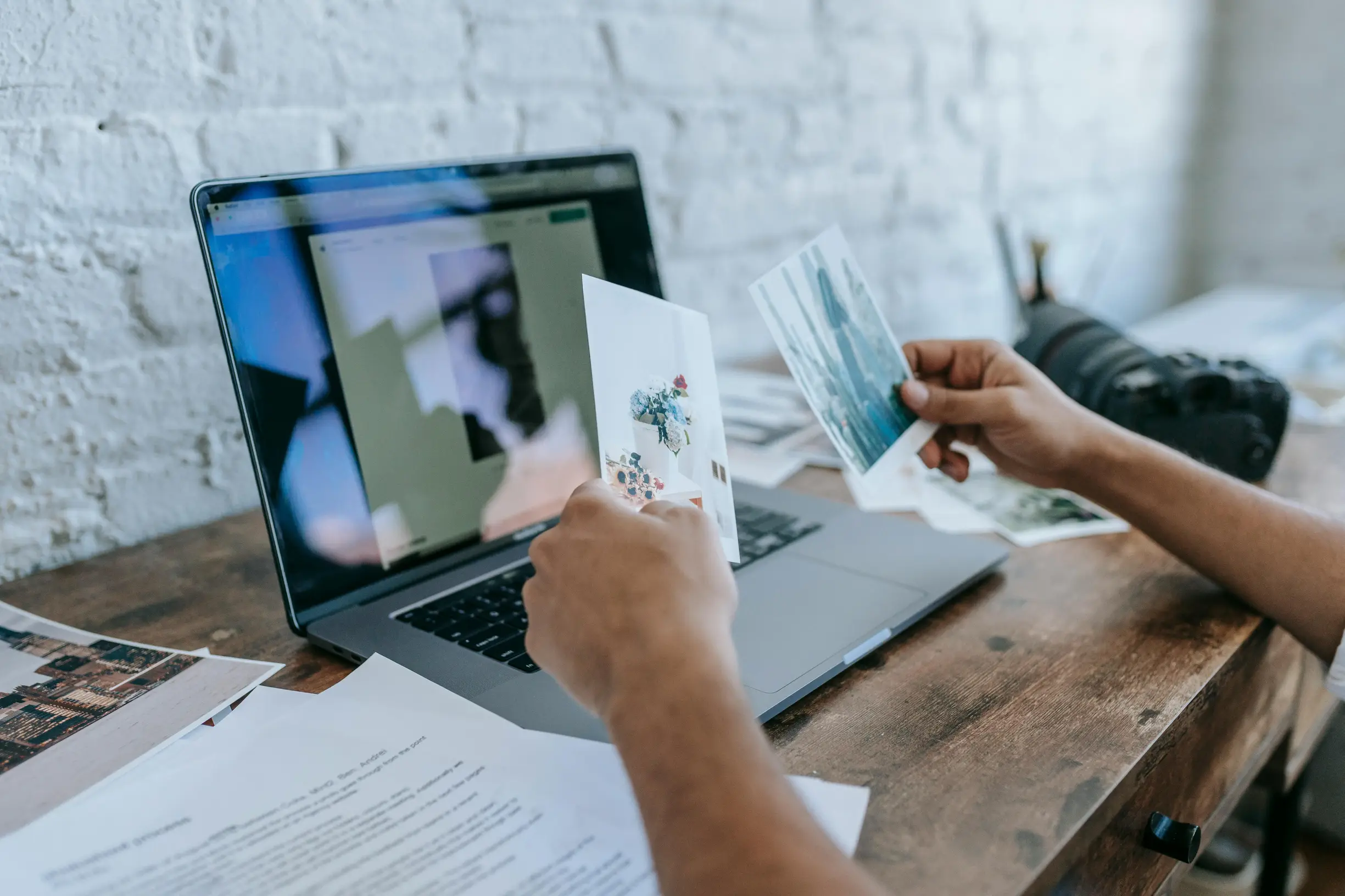 The image shows a person's hands holding several printed photographs above a wooden desk. On the desk is an open laptop displaying a blurry or out-of-focus image on its screen. Papers and documents lie scattered on the desk, and a camera is visible in the background on the right side. The setting seems to be indoors with a white brick wall in the background. The shot is taken from over the shoulder at an eye-level angle, focusing mainly on the hands, photographs, and laptop.