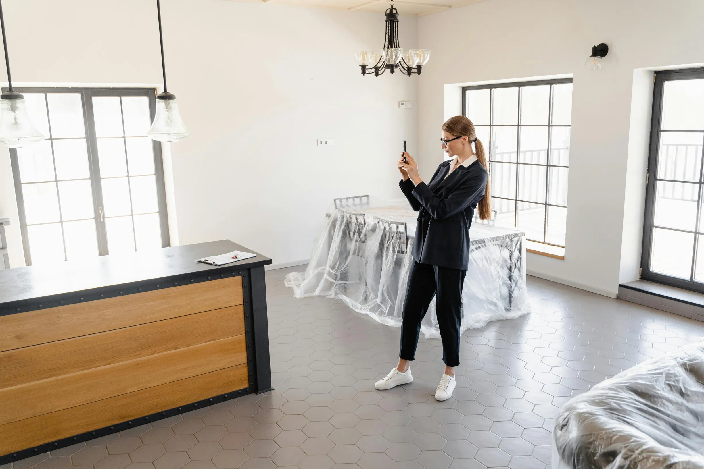 The image shows a young adult woman standing indoors in a room with large windows letting in natural light. The room appears mostly empty, with furniture covered in clear protective plastic sheets, suggesting a recent move or renovation. The floor has a hexagonal tile pattern. The woman is dressed in a black blazer, black pants, white sneakers, and glasses, and is holding a smartphone at eye level, seemingly taking a photo or video. A wooden counter with a black top is visible on the left side, and a small 