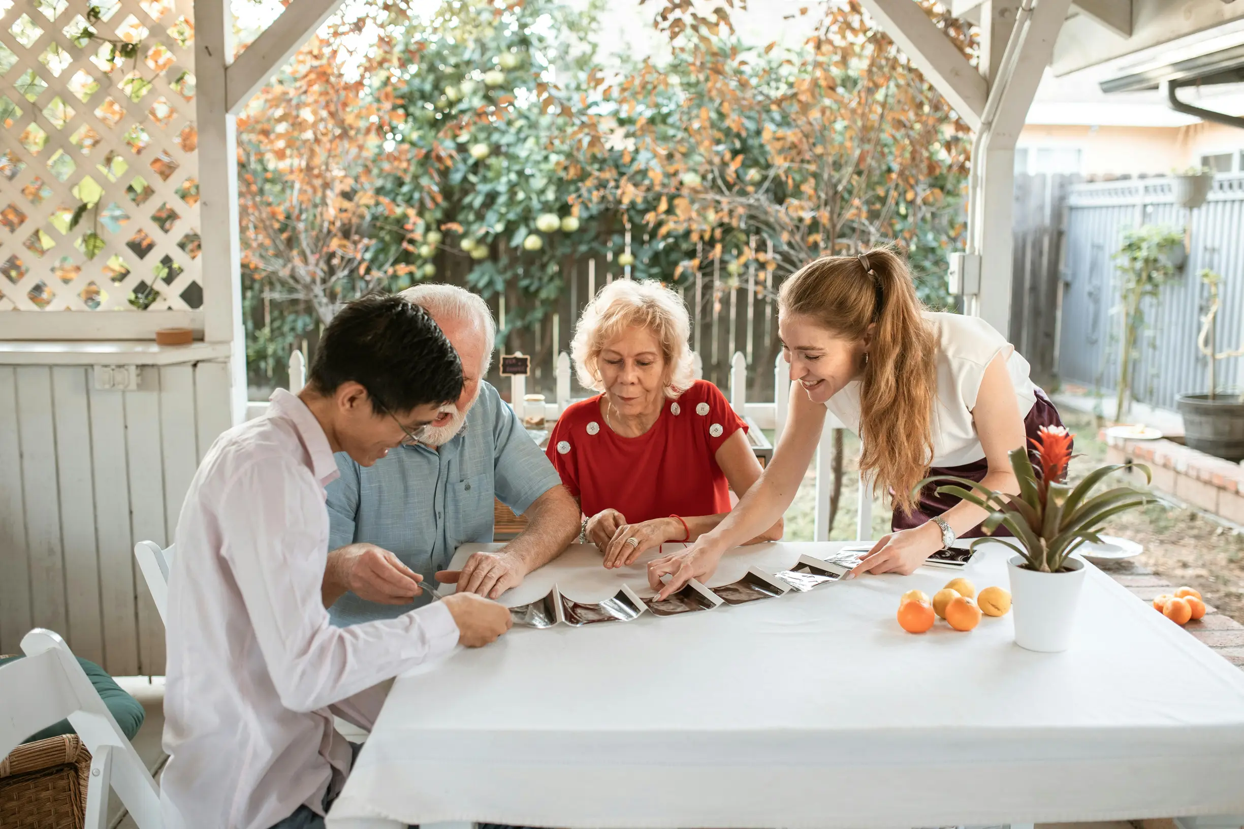 The image shows four people gathered around a white table in an outdoor patio setting under a white pergola. The group consists of an older woman with curly gray hair wearing a red top, an older man in a light blue shirt, a younger woman with long brown hair in a white shirt, and a younger man with short dark hair wearing a white long-sleeve shirt. They are engaged in looking at and sorting through a spread of photographs or cards on the table. The background reveals a garden with green foliage and a wooden