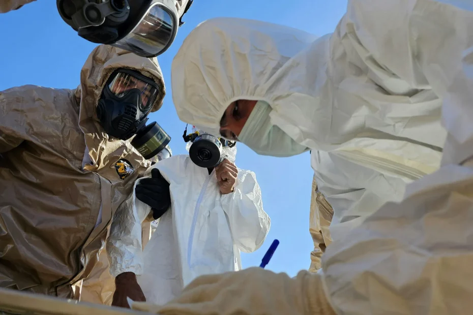 The image shows three people wearing full-body protective suits and respirator masks. One person in the foreground is dressed in a white suit with a hood and mask, bending over and writing with a blue pen. Two other individuals in the background wear tan protective suits and black respirator masks, one facing forward and the other looking slightly downward. The background is a clear blue sky with natural lighting, indicating the scene is outdoors.