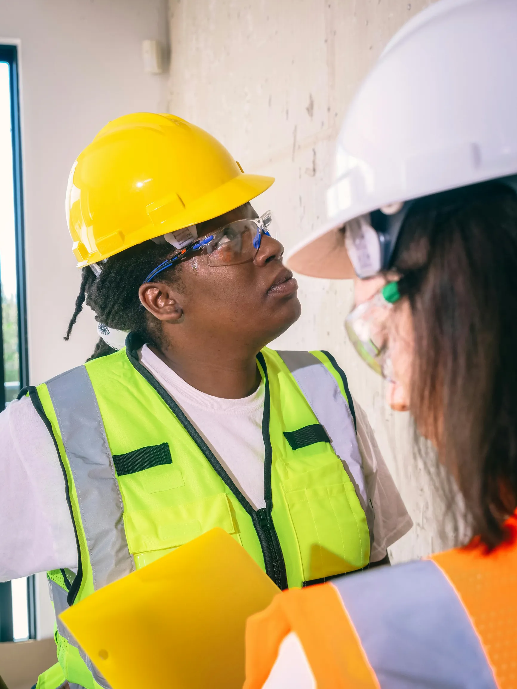 The image shows two construction workers indoors, standing near a wall. The worker on the left is a Black woman wearing clear protective glasses, a yellow hard hat, and a neon yellow safety vest over a white shirt. She holds a yellow clipboard and looks attentively at the wall. The other worker, partially visible from behind, wears a white hard hat, glasses, and an orange safety vest with reflective stripes. The background suggests a construction or renovation site with an unfinished wall and natural light 