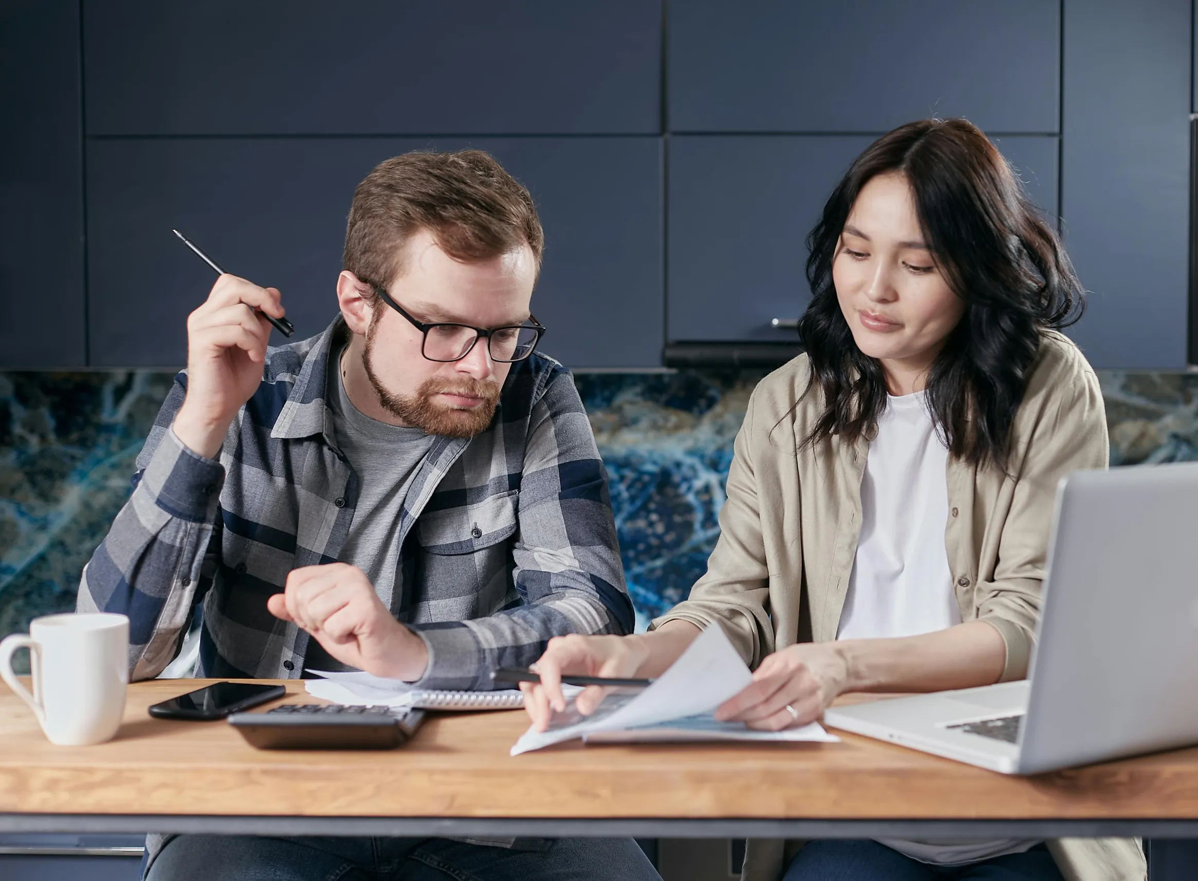 Two adults, one male and one female, are seated at a wooden table indoors, working together. The man has light skin, short brown hair, a beard, and wears glasses, a gray and black plaid shirt over a dark gray t-shirt. The woman has light to medium skin, long black hair, and wears a white shirt under a light beige cardigan. They are focused on paperwork spread on the table alongside a calculator, smartphone, and a white mug. A silver laptop is open in front of the woman. The background shows blue cabinets an